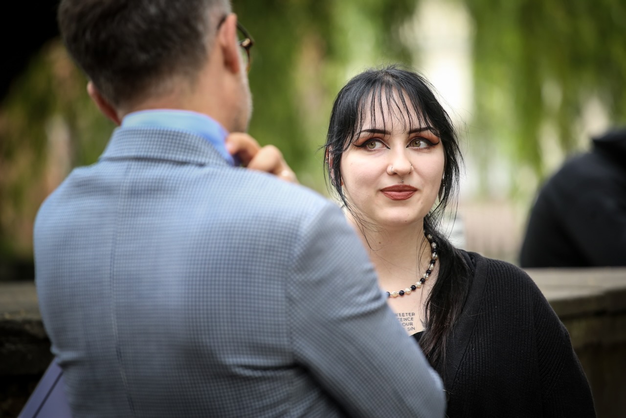 Młoda biała kobieta, w czwarnych włosach, z opadającą na twarz grzywką i mocnym makijażem. Patrzy na mężczyznę w garniturze (którego widać tylko plecy w jasnej marynarce)