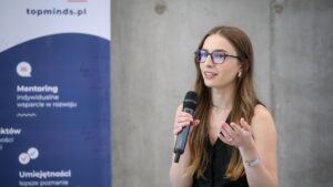 Młoda, biała kobieta w czarnej bluzce odsłaniającej ramiona, w okularach i długich ciemnych włosach, trzymająca mikrofon w prawej ręce. Za nią betonowa ściana i niebieski baner z napisem "mentoring"