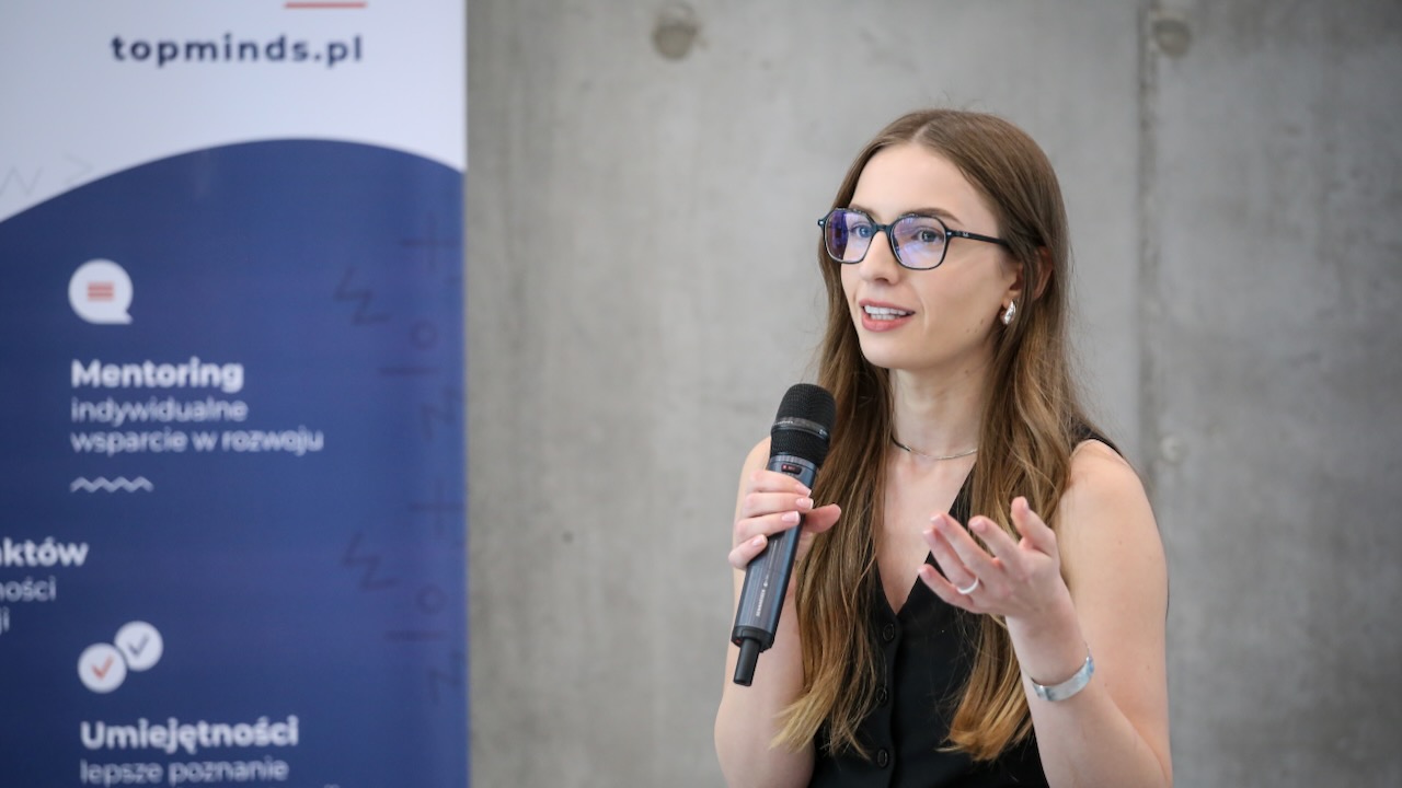 Młoda, biała kobieta w czarnej bluzce odsłaniającej ramiona, w okularach i długich ciemnych włosach, trzymająca mikrofon w prawej ręce. Za nią betonowa ściana i niebieski baner z napisem "mentoring"