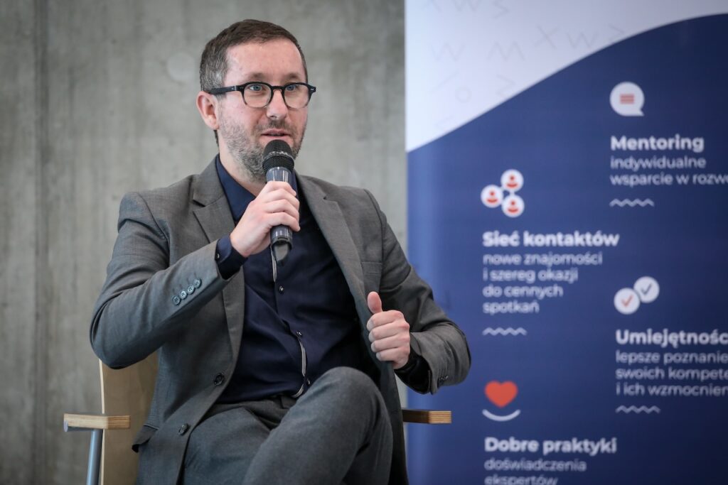 Biały mężczyzna w okularach i garniturze trzyma mikrofon w prawej ręce. Obok niego niebieski baner z hasłami: "mentoring", "sieć kontaktów", "umięjętności", "dobre praktyki".