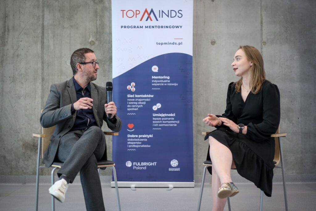 Dwie osoby siedzą na krzesłach patrząc na siebie. Jedna z nich trzyma mikrofon. W tle stoi biało-niebieski baner z logo TopMinds.