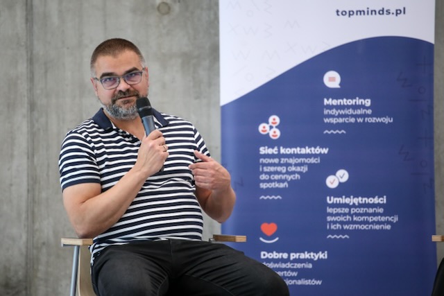 Biały mężczyzna w średnim wieku, w okularach i bluzce w paski trzyma mikrofon w prawej ręce. Obok niego niebieski baner z hasłami: "mentoring", "sieć kontaktów", "umięjętności", "dobre praktyki".