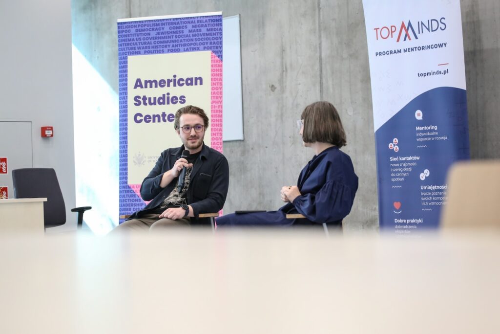 Dwie osoby w czarnych ubraniach siedzą naprzeciwko siebie i rozmawiają. W tle bannery z napisem "American Studies Center" i TopMinds