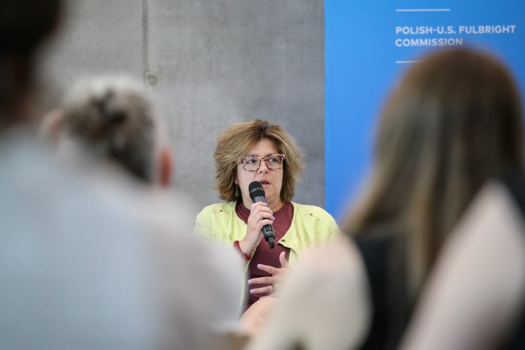 Kobieta w żółtej bluzce i okularach trzyma mikrofon. Na pierwszym planie rozmazane postaci z widowni.