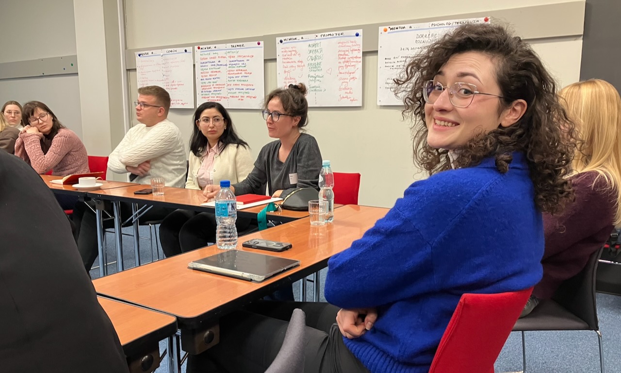 Uśmiechnięta młoda dziewczyna w ciemnych kręconych włosach i okularach siedzi przy stole na szkoleniu