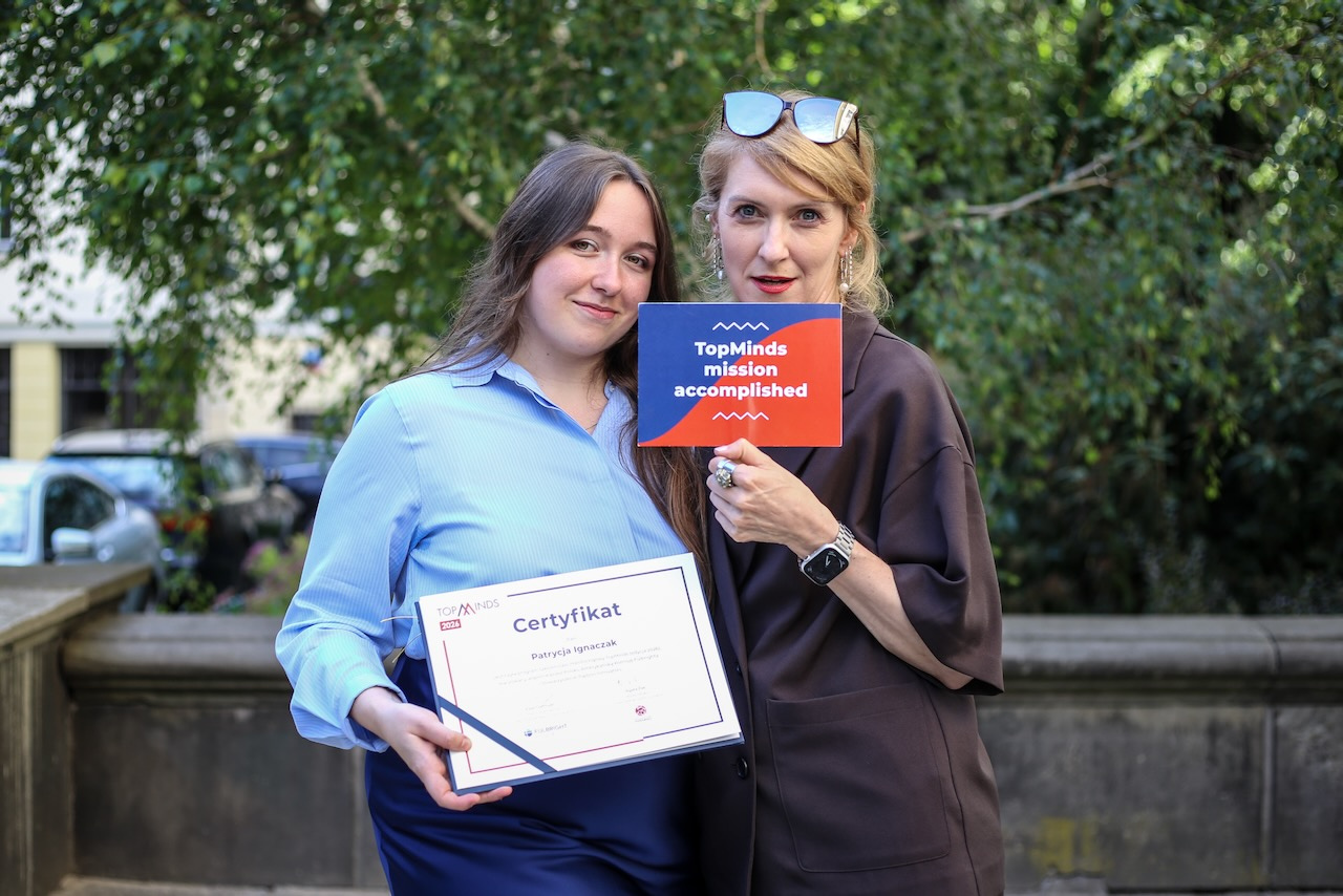 Dwie kobiety na tle zieleni, trzymające dyplom zakończenia programu i tabliczkę z napisem "TopMinds mission accomplished"