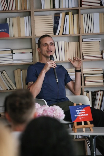 Mężczyzna w niebieskim t-shircie na tle książek, mówiący do mikrofonu i gestykulujący