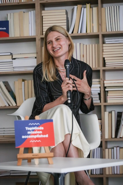 Uśmiechnięta kobieta w długich blond włosach i czarnej bluzce siedząca na tle regału z ksiażkami, trzymająca mikrofon. Przed nią czerwono-niebieska tabliczka z napisem "TopMinds mission accomplished"