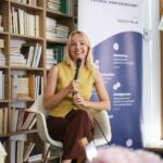 Młoda kobieta w blond włosach i żółtej bluzce, siedząca na tle biało-niebieskiego baneru z napisem TopMinds, mówiąca do mikrofonu.