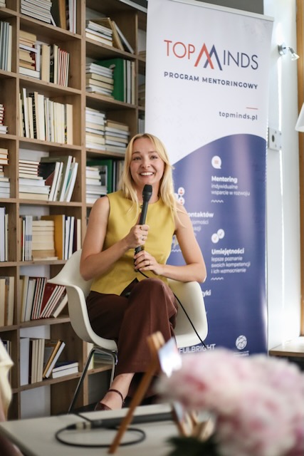 Młoda kobieta w blond włosach i żółtej bluzce, siedząca na tle biało-niebieskiego baneru z napisem TopMinds, mówiąca do mikrofonu.